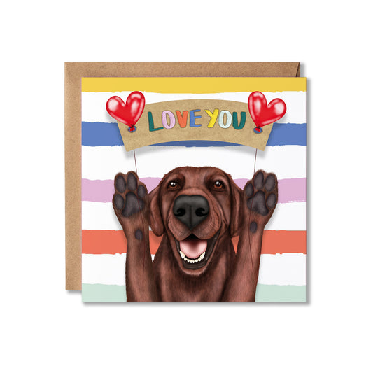 A square greeting card with a brown envelope behind it, featuring an illustration of a smiling brown dog holding red heart-shaped balloons that spell 'LOVE YOU' on a tan banner. The card has horizontal pastel stripes in blue, pink, orange, and mint, with a yellow band at the top.