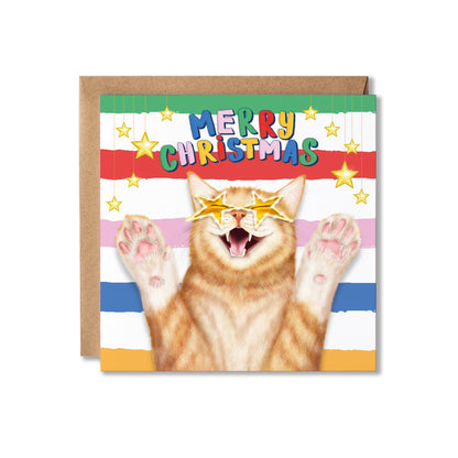 A Christmas greeting card featuring a ginger cat with star-shaped sunglasses and raised paws, with 'MERRY CHRISTMAS' in colourful letters above. The card is shown with a brown kraft envelope behind it on a white background.