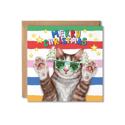 A square Christmas greeting card with a brown kraft envelope behind it. The card features a tabby cat wearing green Christmas tree sunglasses, with colourful striped bands and the text “MERRY CHRISTMAS” across the top.