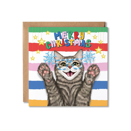 A Christmas greeting card featuring a tabby cat wearing blue snowflake glasses, with colourful striped bands and the text 'MERRY CHRISTMAS' at the top. A brown kraft envelope is partially visible behind the card on a white background.