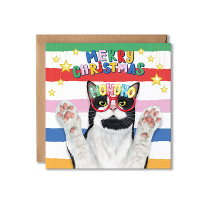 A square Christmas greeting card with a brown kraft envelope behind it. The card features a black-and-white cat wearing red glasses with the word "HOHOHO" on the glasses, set against multicolored horizontal stripes and the text "MERRY CHRISTMAS" with hanging stars.