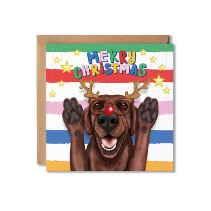 A square Christmas greeting card featuring a brown Labrador dog dressed as a reindeer with antler and red nose glasses and raised paws, with the text 'MERRY CHRISTMAS' above. The card is shown with a brown kraft envelope.