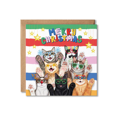 A Christmas greeting card featuring seven cats wearing Christmas sunglasses, with the text 'MERRY CHRISTMAS' at the top of a colourful striped background and hanging stars. A brown kraft envelope is placed behind the card on a plain white background.