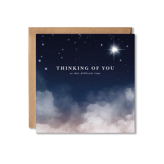 A square greeting card with a kraft paper envelope, featuring a night sky design with stars, a bright starburst, and soft clouds at the bottom. The card reads 'THINKING OF YOU at this difficult time'.
