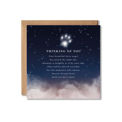 A square greeting card with a kraft envelope behind it, featuring a night sky design with stars, a glowing paw print constellation, and soft pink clouds at the bottom. White text on the card reads, 'THINKING OF YOU' followed by a comforting message.