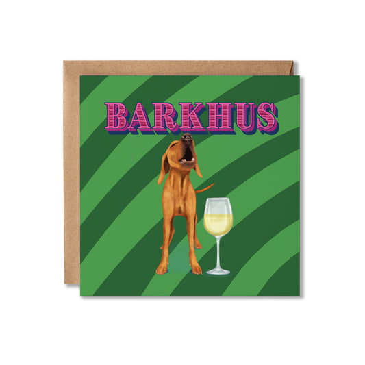 A square greeting card with a kraft envelope behind it, featuring a brown dog standing next to a wine glass on a green striped background, with the word 'BARKHUS' in pink outlined letters at the top.
