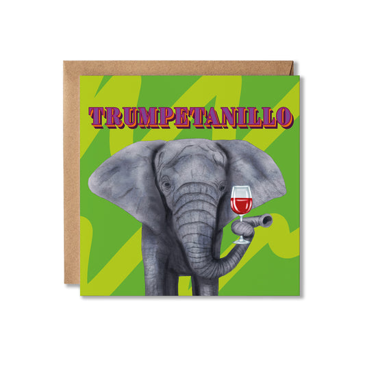 A square greeting card with a kraft envelope behind it, featuring an illustration of a gray elephant holding a glass of red wine against a bright green patterned background. The card reads 'TRUMPETANILLO' in stylized pink and purple lettering.