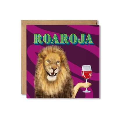 A square greeting card with a kraft envelope behind it, featuring a lion illustration holding a glass of red wine against a purple striped background, with the word 'ROAROJA' in green outlined lettering at the top.