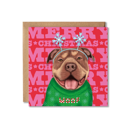 A square Christmas greeting card with a kraft envelope behind it. The card features an illustrated pitbull-type dog wearing a green knit sweater that says 'woof' and blue snowflake antlers, set against a pink background with repeated 'MERRY CHRISTMAS' text.