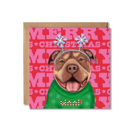 A square Christmas greeting card with a kraft envelope behind it. The card features an illustrated pitbull-type dog wearing a green knit sweater that says 'woof' and blue snowflake antlers, set against a pink background with repeated 'MERRY CHRISTMAS' text.