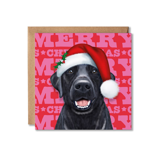 A square Christmas greeting card featuring a black dog wearing a red Santa hat with a white pom-pom, set against a pink patterned background with the word 'MERRY CHRISTMAS' repeated. A brown kraft envelope is placed behind the card on a white background.