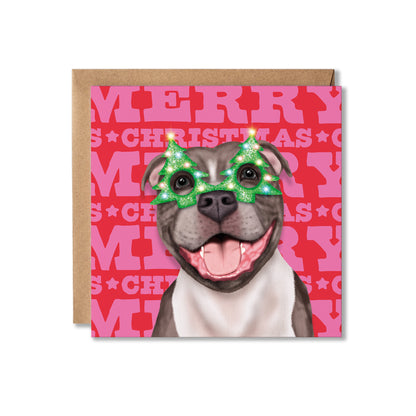 A square Christmas greeting card featuring an illustration of a Staffordshire bull terrier dog wearing green glittery Christmas tree-shaped sunglasses, set against a pink and red background with 'MERRY CHRISTMAS' text, shown with a brown kraft envelope behind it on a white background.