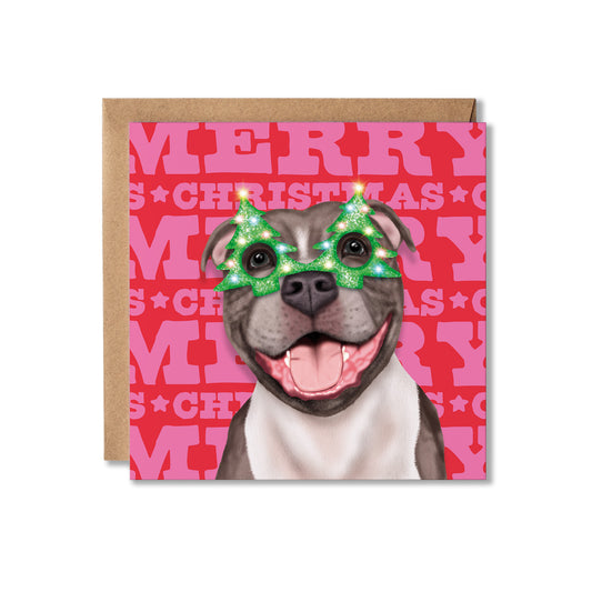 A square Christmas greeting card featuring an illustration of a Staffordshire bull terrier dog wearing green glittery Christmas tree-shaped sunglasses, set against a pink and red background with 'MERRY CHRISTMAS' text, shown with a brown kraft envelope behind it on a white background.