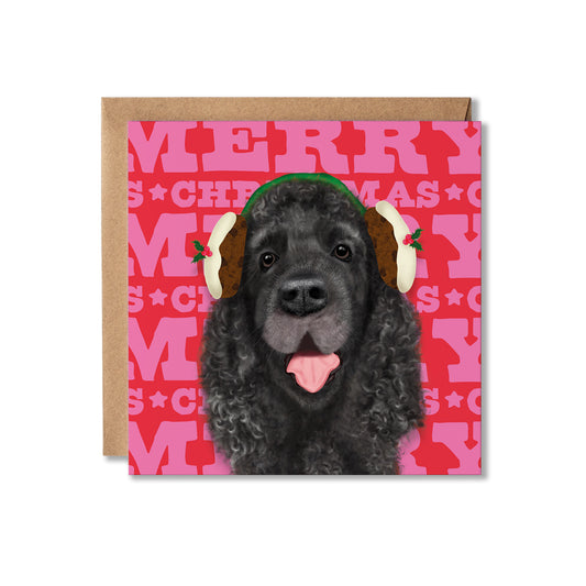 A square Christmas greeting card featuring a black dog wearing cream and brown holiday earmuffs against a pink and red patterned background with the words 'MERRY CHRISTMAS.' A kraft paper envelope is placed behind the card on a white background.