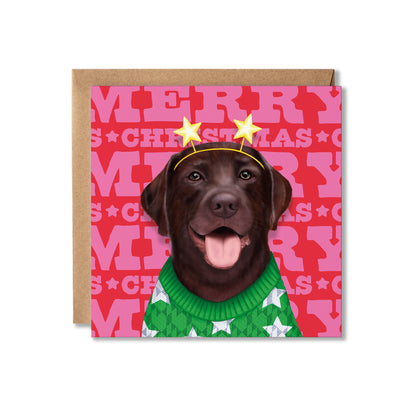 A square Christmas greeting card featuring a chocolate Labrador retriever wearing a green knit sweater with white stars and a yellow star headband, set against a pink and red patterned background with 'MERRY CHRISTMAS' text. A brown kraft envelope is partially visible behind the card.