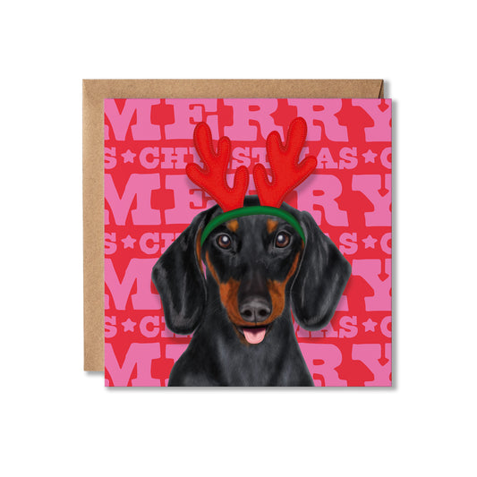 A square Christmas greeting card featuring a black and tan dachshund wearing red reindeer antlers and a green headband, set against a pink and red Merry Christmas background. A brown kraft envelope is partially visible behind the card on a white background.