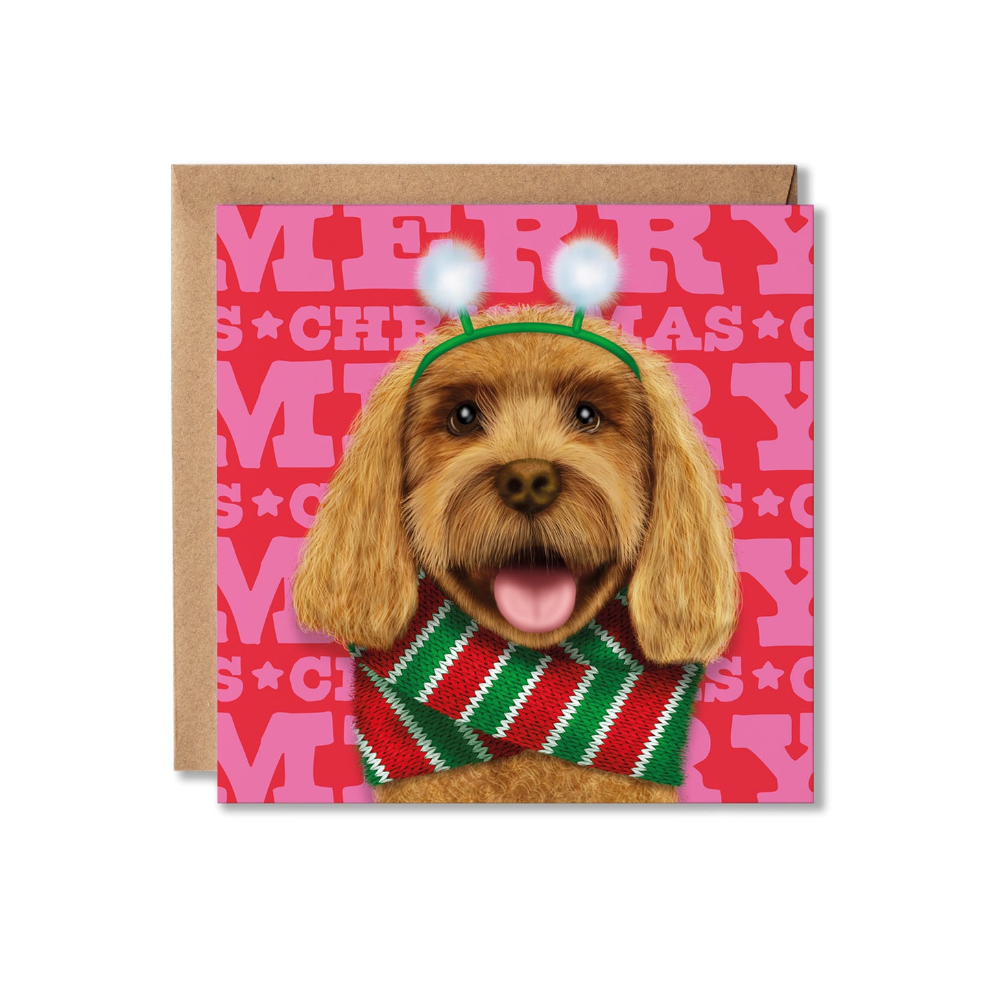 A square Christmas greeting card featuring a brown dog wearing a green antler headband with white pom-poms and a red-and-green striped knit sweater, set against a pink holiday patterned background. A brown kraft envelope is partially visible behind the card on a white studio background.