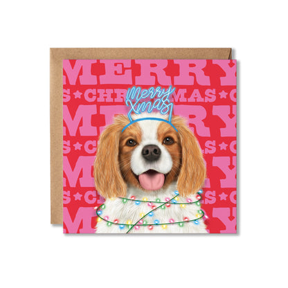 A square Christmas greeting card featuring a Cavalier King Charles Spaniel dog wearing a blue 'Merry Xmas' hat and colorful holiday lights, set against a pink patterned background with the word 'MERRY' repeated. A brown kraft envelope is placed behind the card on a white background.
