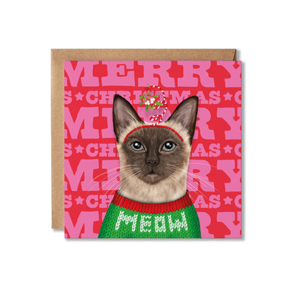A square Christmas greeting card featuring a Siamese cat wearing a green knit sweater that says 'MEOW' and a red hat with a candy cane, set against a pink background with 'MERRY CHRISTMAS' text, shown with a brown kraft envelope behind it on a white background.