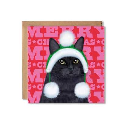 A square Christmas greeting card featuring a black cat wearing a green and white Santa hat with white pom-poms, set against a pink patterned background with the word 'MERRY' repeated. A brown kraft envelope is partially visible behind the card on a white background.