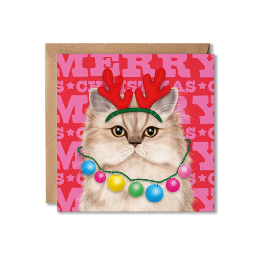 A square Christmas greeting card featuring a fluffy cream-colored cat wearing red reindeer antlers and a colorful string of holiday lights, set against a pink patterned background with the word 'MERRY' repeated. A brown kraft envelope is partially visible behind the card on a white background.