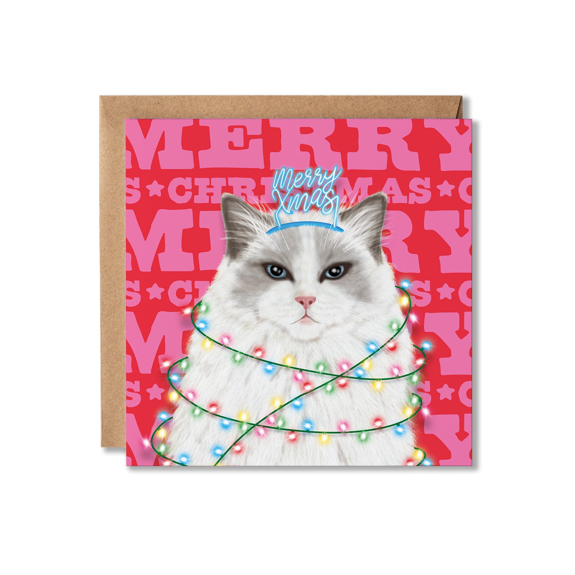 A square Christmas greeting card featuring a fluffy white cat with blue eyes, wrapped in multicoloured holiday lights and wearing a blue crown, set against a red background with pink 'MERRY CHRISTMAS' lettering. A brown kraft envelope is placed behind the card on a white background.