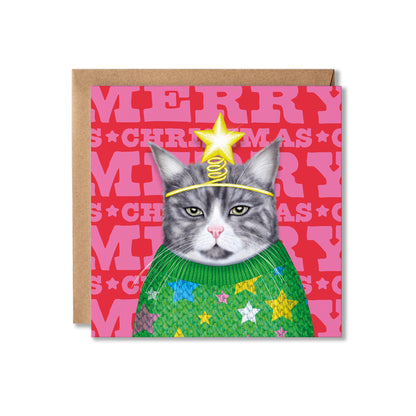 A square Christmas greeting card featuring a gray cat wearing a green knit sweater with colorful stars and a gold star-and-coil headband, set against a pink patterned background. A brown kraft envelope is placed behind the card on a white studio background.