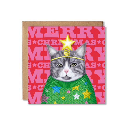 A square Christmas greeting card featuring a gray cat wearing a green knit sweater with colorful stars and a gold star-and-coil headband, set against a pink patterned background. A brown kraft envelope is placed behind the card on a white studio background.