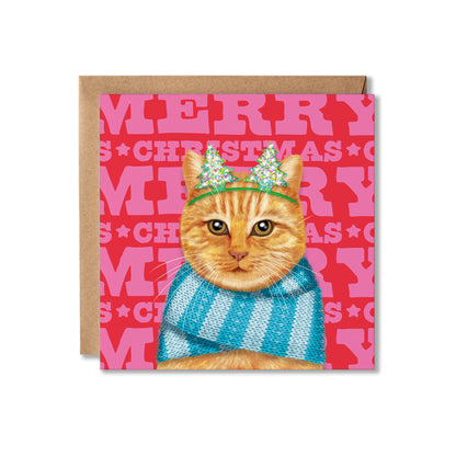 A square Christmas greeting card featuring an orange tabby cat wearing a green glittery crown and a blue striped knit scarf, set against a red and pink background with the words 'MERRY CHRISTMAS'. A brown kraft envelope is placed behind the card on a white background.