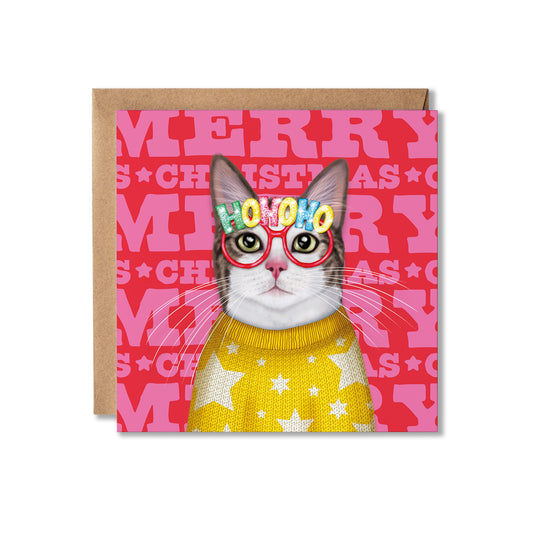 A square Christmas greeting card featuring a gray and white cat wearing red-framed glasses with the word 'HONOKO' in colorful letters and a yellow sweater with white stars, set against a pink background with repeated 'MERRY CHRISTMAS' text. A brown kraft envelope is placed behind the card on a white background.