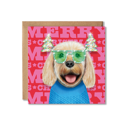 A square Christmas greeting card featuring a dog illustration wearing green glittery sunglasses and a blue sweater, with pink 'MERRY CHRISTMAS' lettering in the background. A brown kraft envelope is placed behind the card on a white studio background.